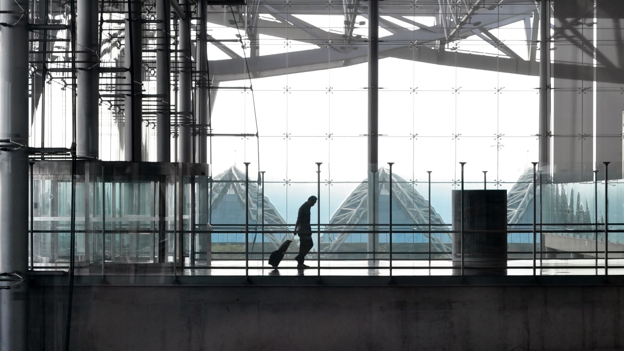 Silhouetted traveler wheeling a suitcase along a glass-walled airport terminal walkway