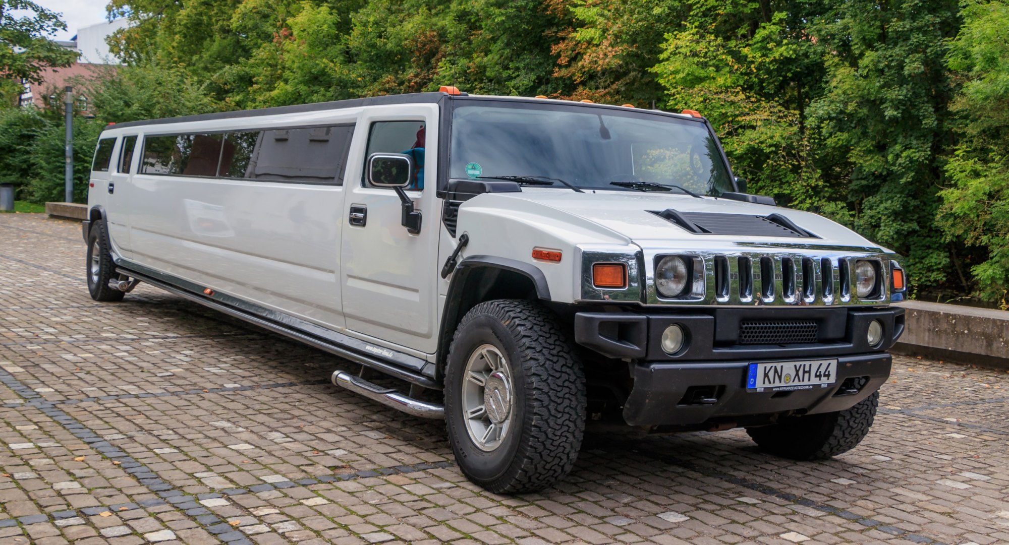 White stretch Hummer H2 limousine parked on a cobblestone lot in front of trees