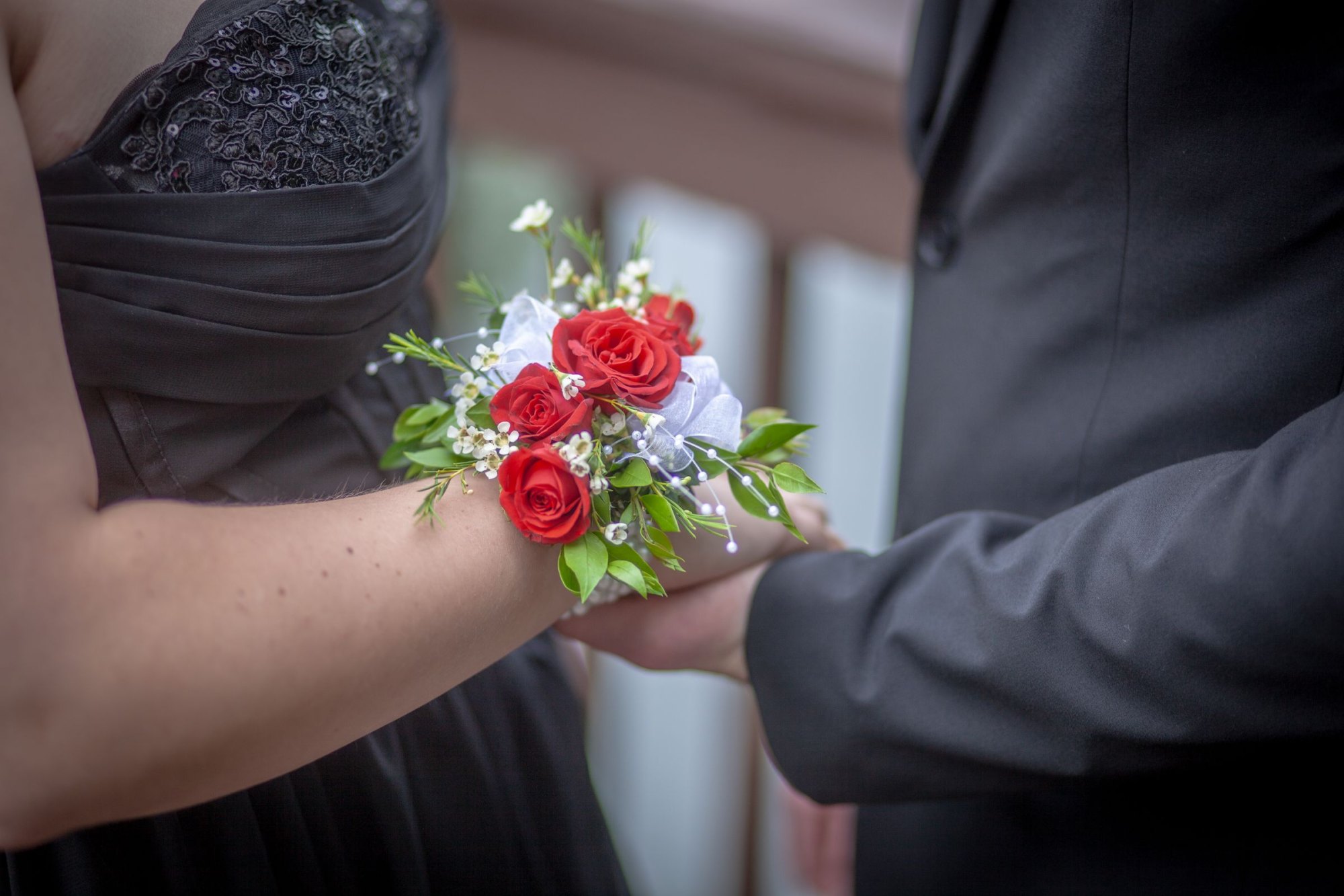 Red rose wrist corsage on a woman in a black gown holding her suited partner’s hand