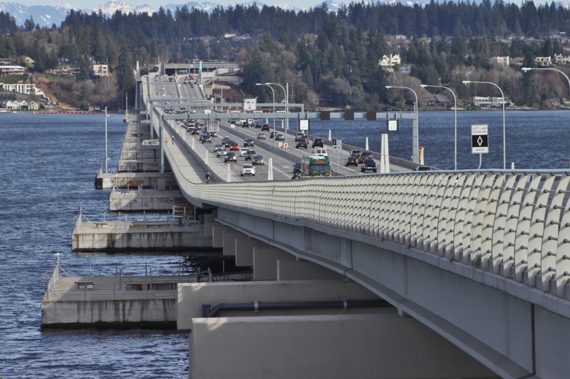 Traffic crossing a floating highway bridge on concrete pontoons over Lake Washington toward wooded hills